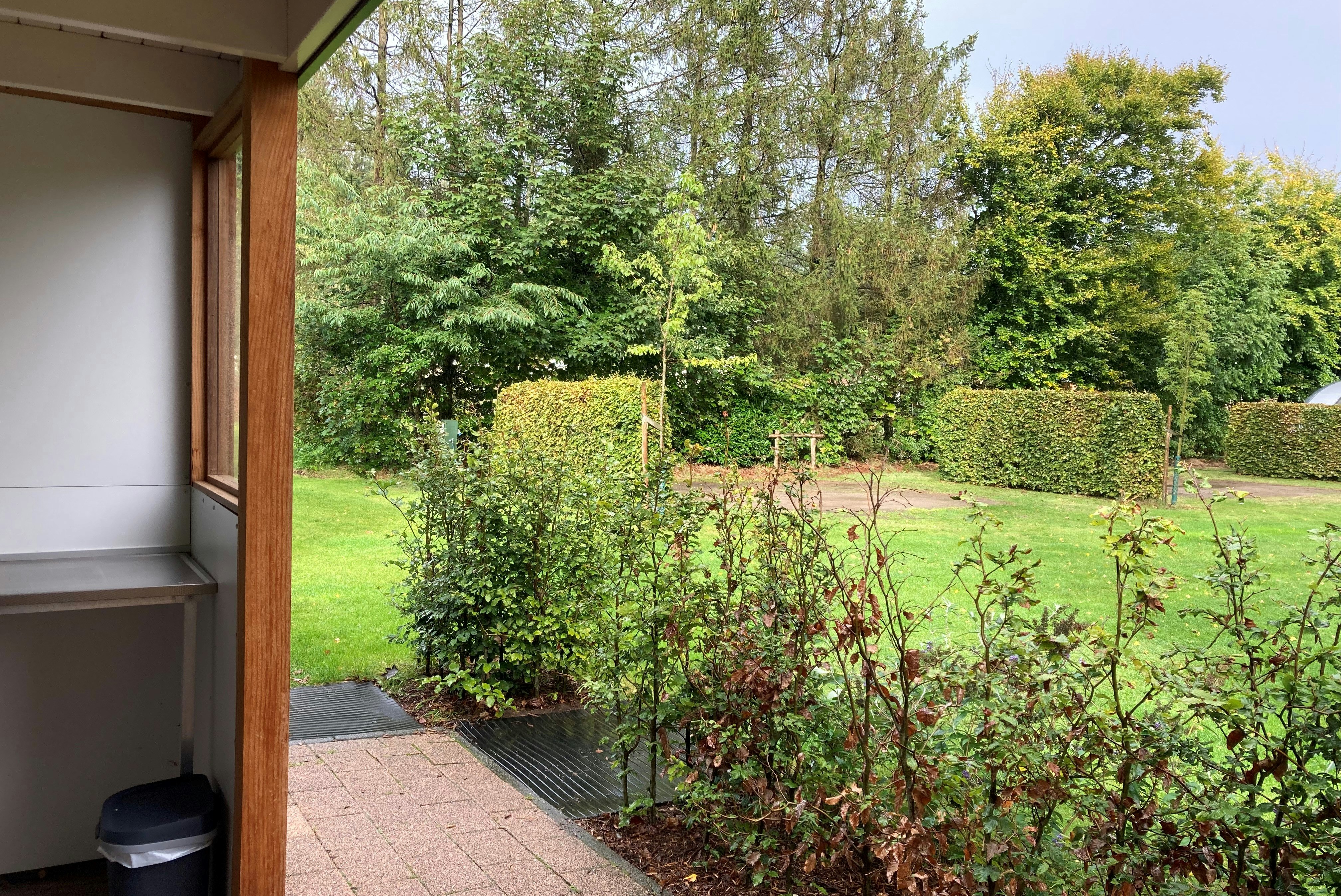 Terrasse am Campingplatz mit Blick auf gepflegten Garten, Hecken und Blumenbeete neben einem Holzrahmen am Gebäude.