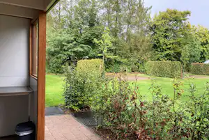 Terrasse am Campingplatz mit Blick auf gepflegten Garten, Hecken und Blumenbeete neben einem Holzrahmen am Gebäude.