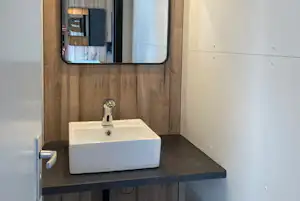 Waschbecken in einem modernen Sanitärmodul mit Holzverkleidung, Spiegel und Wandbeleuchtung.