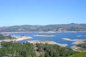 Blick auf eine Bucht mit Wasser, Küste und Hügeln, im Vordergrund Bäume und ein Weg am Campingplatz.