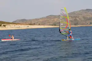 Windsurfer auf dem See mit Segel, daneben ein SUP mit Paddler vor sandigem Ufer und Hügeln.