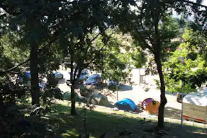 Zelte auf einem Campingplatz zwischen Bäumen, im Hintergrund Autos und ein Wohnwagen auf dem Stellplatz.