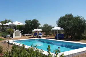 Schwimmbecken mit blauem Wasser, weiße Sonnenschirme und Campingzelte im Hintergrund, umgeben von Bäumen und Wegen.