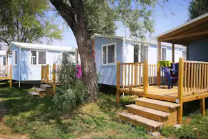 Holzterrasse mit Sitzgelegenheiten vor einem weißen Mobilheim, umgeben von Bäumen und Rasen auf dem Campingplatz.