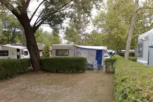 Stellplatz mit Wohnwagen und Zelt, Sitzbank und Tisch im Grünen zwischen Hecken und Bäumen.