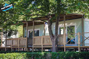 Bungalow auf dem Campingplatz mit Holzveranda, Geländer und großen Fenstern im Grünen, umgeben von Bäumen.