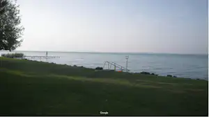 Grüner Campingplatz am Meer mit Blick auf Wasser, Steg und Treppe am Ufer, im Hintergrund Bäume und Himmel.