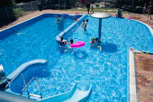 Kinder im großen Camping-Schwimmbad mit blauem Wasser, Schwimmreifen und Spielzeugbooten am Beckenrand.