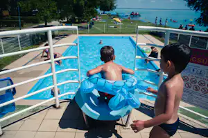 Kinderpool am Campingplatz mit zwei Kindern im Wasser, Sonnenschirm und Blick auf den See im Hintergrund.