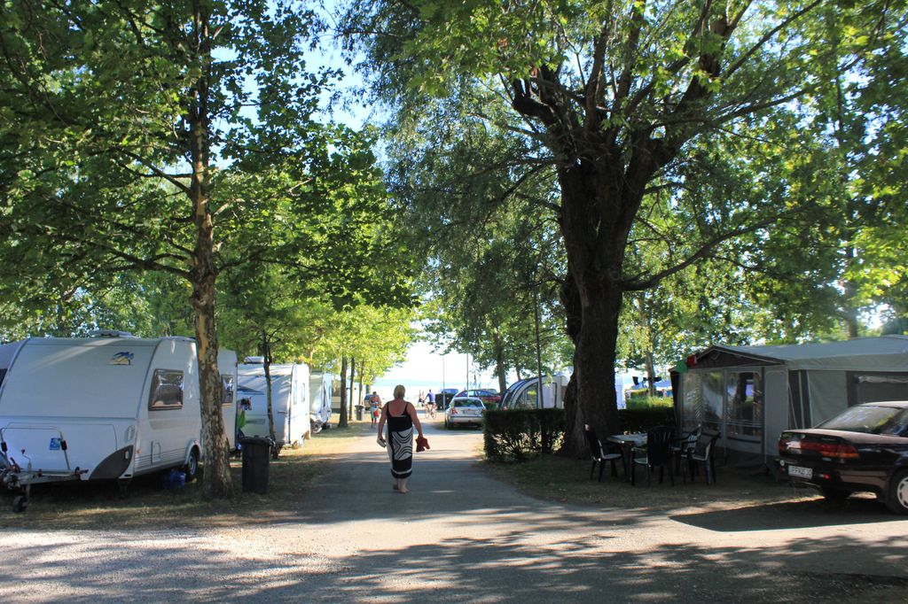 Campingplatz mit Wohnwagen und Zelt zwischen Bäumen, gepflasterter Weg, Autos und Sitzgruppe am Stellplatz.