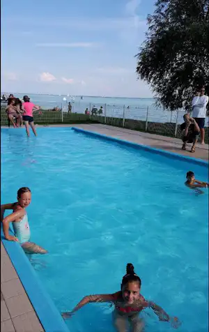 Schwimmbad am Campingplatz mit Blick auf den See, Kinder im Wasser und Menschen am Beckenrand.
