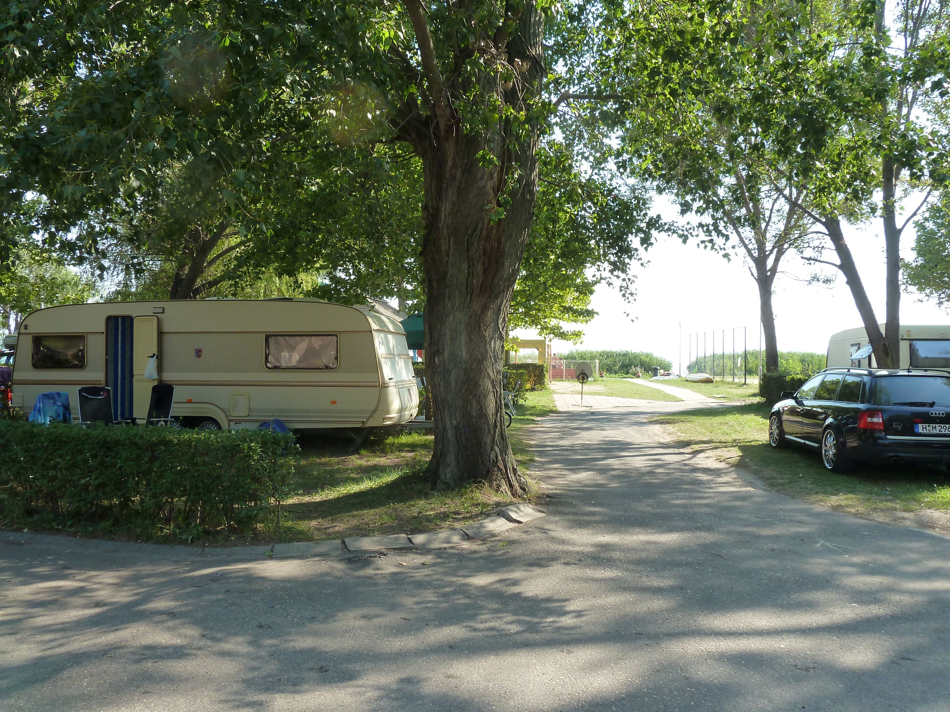 Wohnwagen auf einem Campingplatz-Stellplatz zwischen Bäumen, daneben ein Auto am Weg.