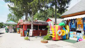Pizza-Stand und Camping-Shop mit bunten Dekoartikeln, Verkaufsregalen und Sitzbank am Weg, neben Bäumen und einem Gebäude.