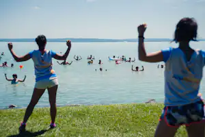 Zwei Personen am Seeufer, im Hintergrund schwimmen Menschen im Wasser und liegen Boote auf dem See.