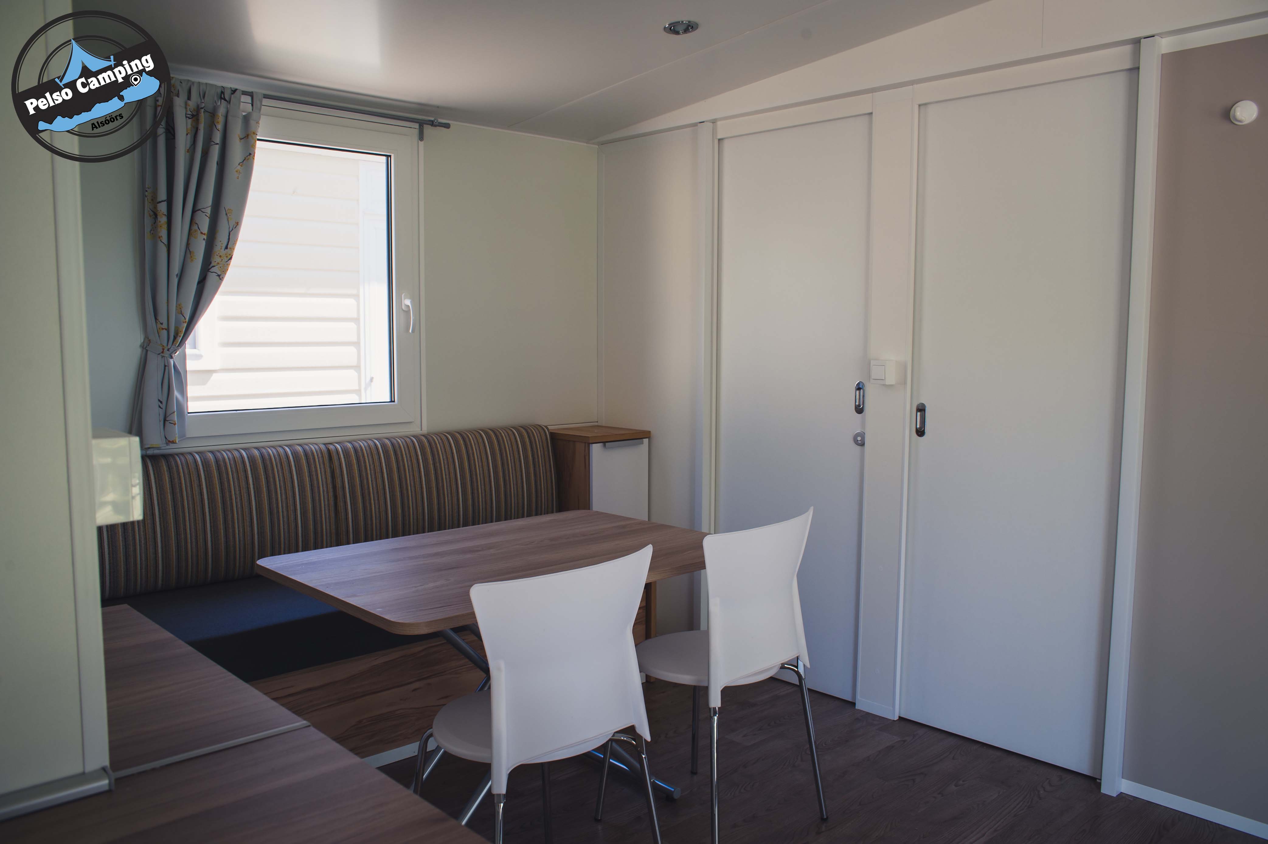 Innenansicht eines Wohnwagens mit Essbereich: Tisch und Stühle, Sitzbank, Schranktüren und Fenster mit Vorhang.