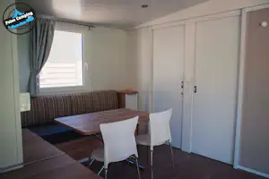 Innenansicht eines Wohnwagens mit Essbereich: Tisch und Stühle, Sitzbank, Schranktüren und Fenster mit Vorhang.