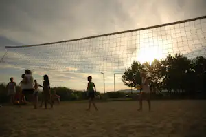 Volleyballfeld auf dem Campingplatz mit Netz, Spielern im Hintergrund und Abendhimmel über Bäumen und Wiese.