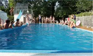 Schwimmbecken mit blauem Wasser, Kinder und Erwachsene am Beckenrand, Sonnenschirme und Bäume im Hintergrund.