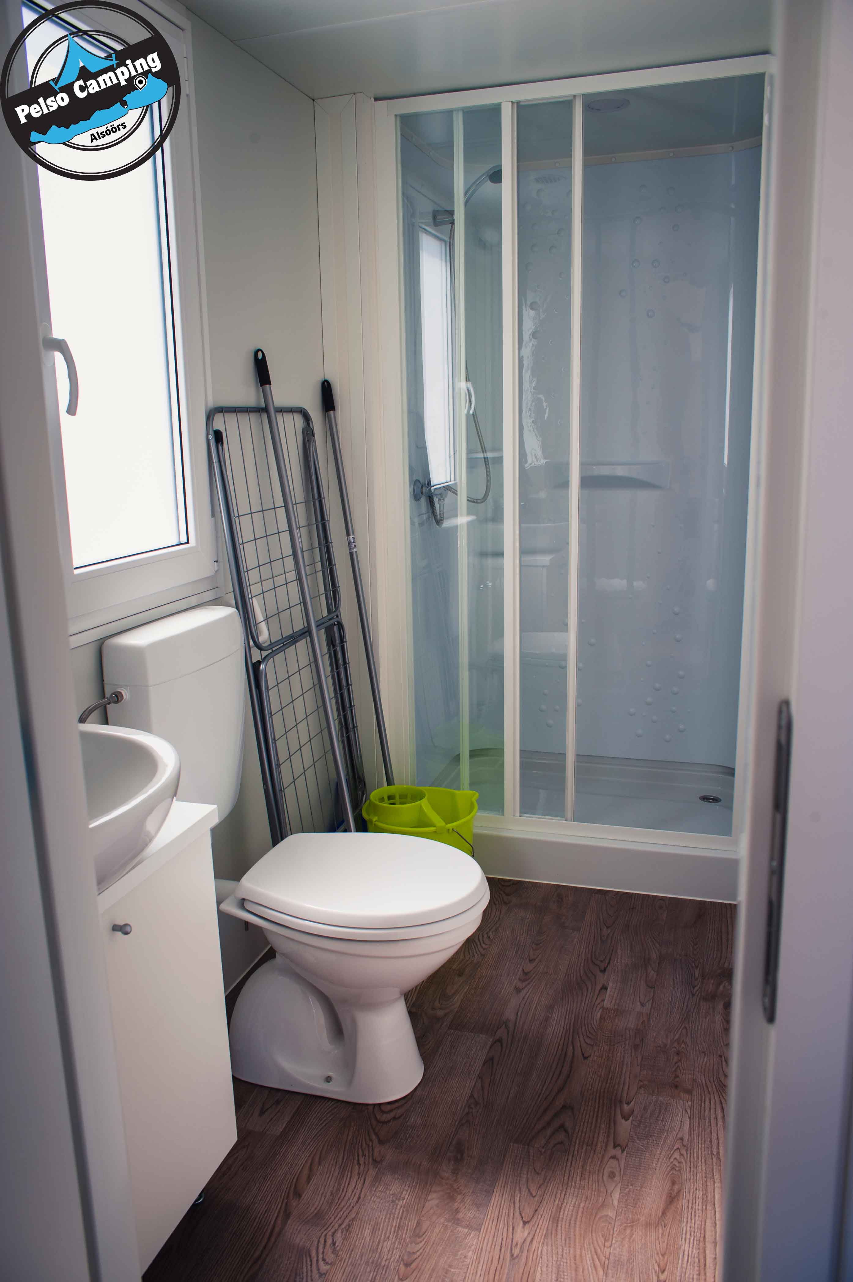 Modernes Badezimmer mit Toilette, Waschbecken und Duschkabine mit Glastür, Boden aus Holzoptik und grünem Eimer.