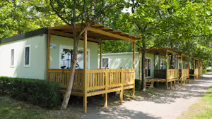 Reihen von Bungalows mit Holzterrassen und Wegen im Grünen, Bäume im Vordergrund und weiße Fassaden am Campingplatz.