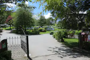 Campingplatz-Einfahrt mit Kiesweg, Metallzaun und vielen Stellplätzen mit Wohnmobilen und Bäumen im grünen Gelände.