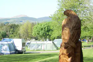 Holzfigur auf dem Campingplatz vor Zelten, Wohnwagen und Bäumen mit Blick auf grüne Hügel im Hintergrund.