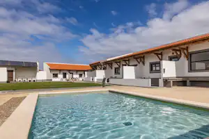 Schwimmbecken mit klarem Wasser vor weißen Ferienhäusern mit Terrassen und orangefarbenen Dächern, im Hintergrund ein blauer Himmel.