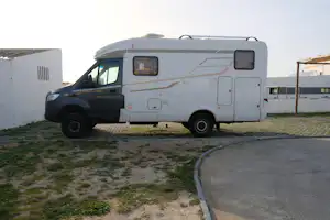 Wohnmobil auf einem Stellplatz mit gepflastertem Boden, neben einem weißen Sanitärgebäude und einem überdachten Bereich.