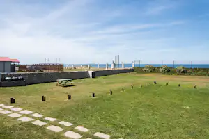 Rasenfläche mit Picknicktisch und niedrigen Holzpfosten, Blick auf das Meer und einen Zaun am Campingplatz.