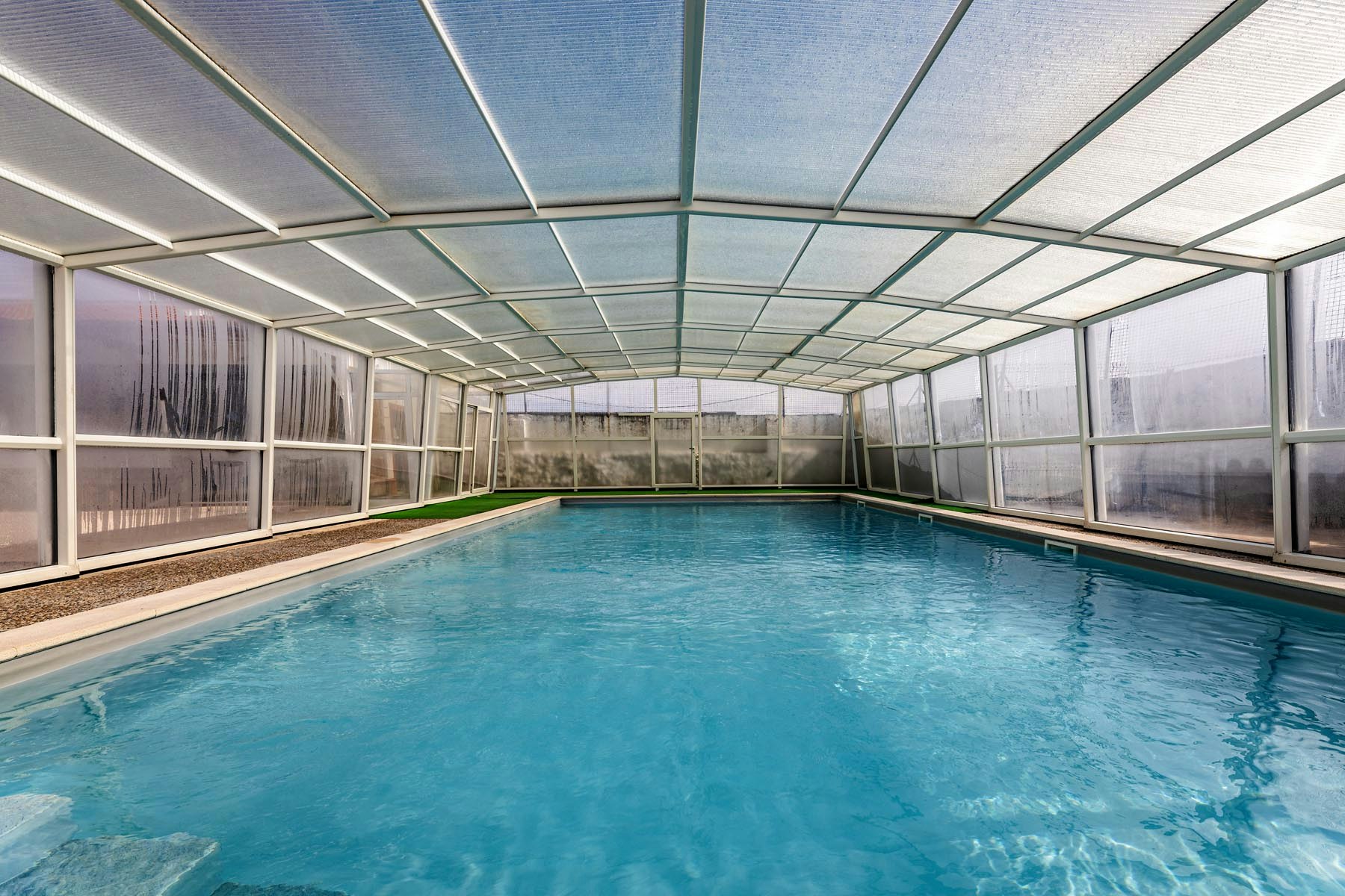 Innenpool mit blauem Wasser unter Glasdach, Seitenwände aus transparenten Paneelen und Metallrahmen, Blick auf das Becken.