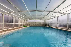 Innenpool mit blauem Wasser unter Glasdach, Seitenwände aus transparenten Paneelen und Metallrahmen, Blick auf das Becken.