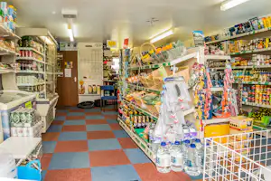 Regale mit Getränken und Lebensmitteln im Camping-Shop, Flaschen und Konserven auf mehreren Ebenen, Kassenbereich im Hintergrund.