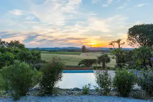 Blick auf den Pool mit Gartenpflanzen und weitem Grünland bei Sonnenuntergang, umgeben von Bäumen und Hügeln.