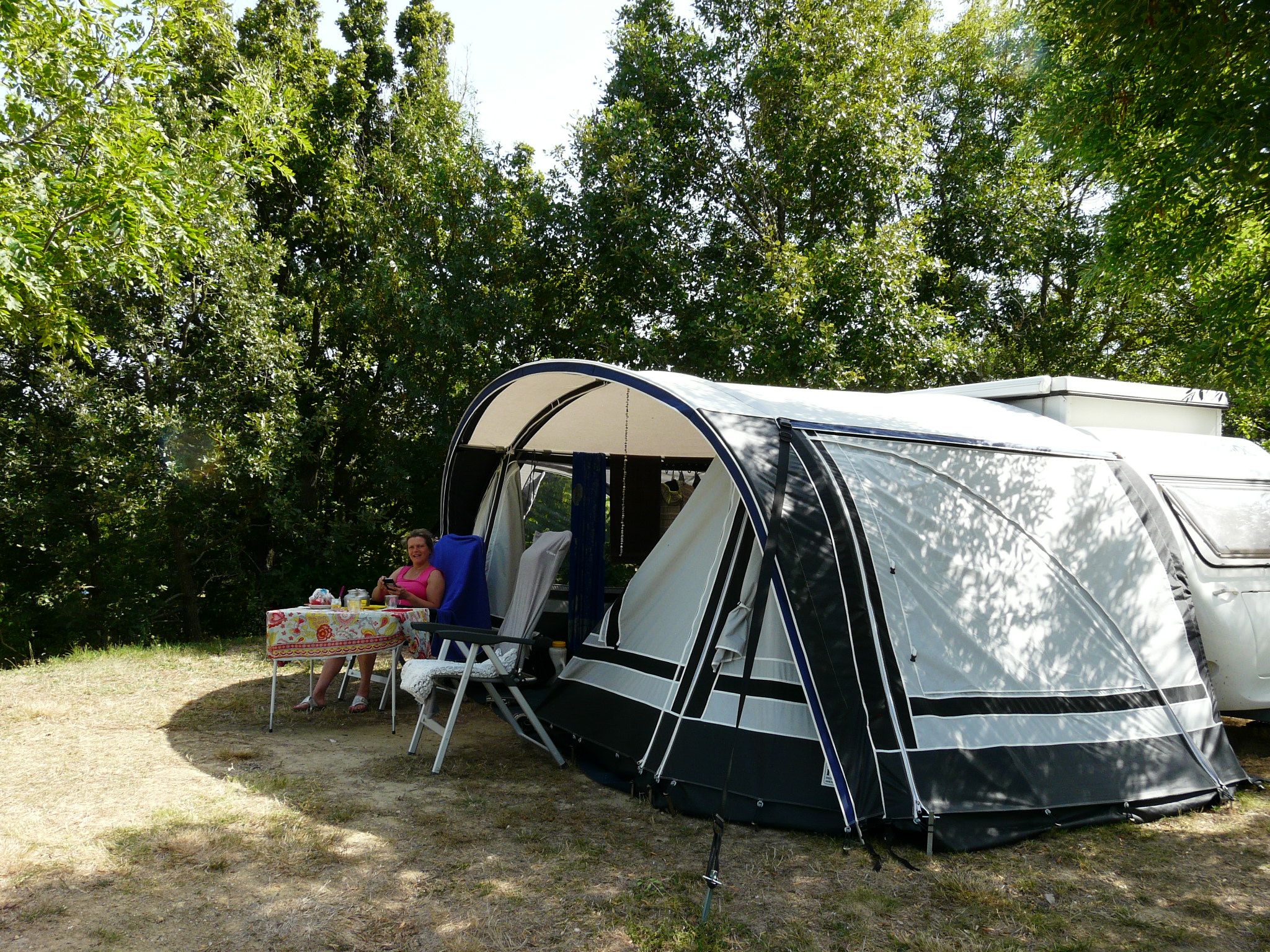 Wohnwagen mit Vorzelt und Campingstuhl am Stellplatz, daneben Tisch, umgeben von Bäumen im Grünen.