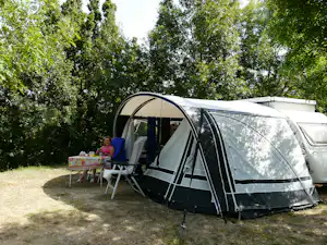 Wohnwagen mit Vorzelt und Campingstuhl am Stellplatz, daneben Tisch, umgeben von Bäumen im Grünen.