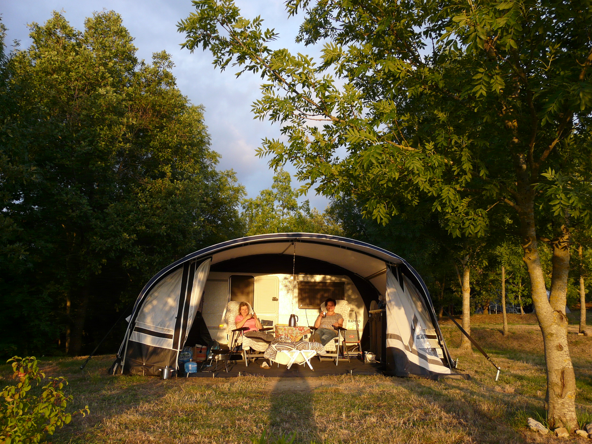 Campingzelt mit offener Vorderseite, Tisch und Stühlen im Inneren, umgeben von Bäumen und Rasen im Grünen.
