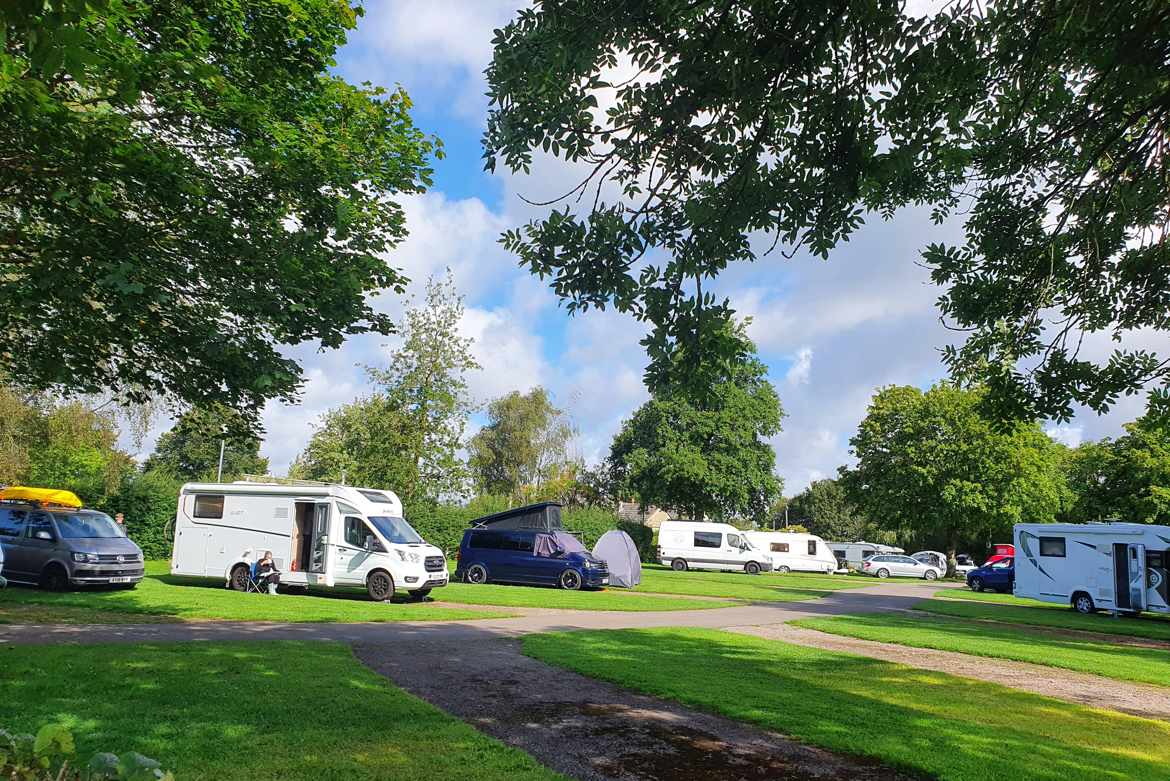 Wohnmobile und Campingbusse auf einer Wiese, umgeben von Bäumen, mit Stellplätzen und Zufahrtsweg im Hintergrund.