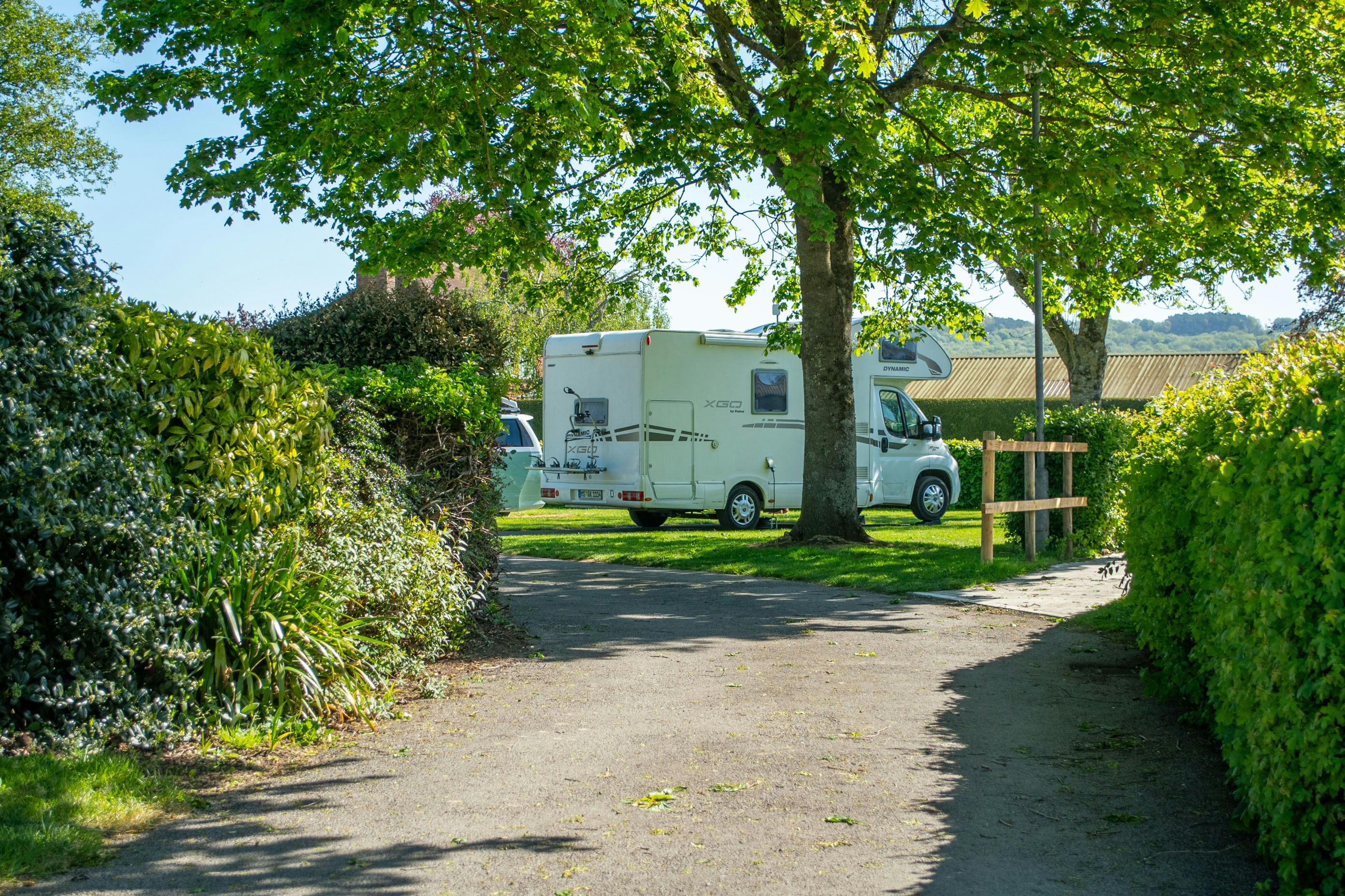 Wohnmobil auf einem Campingplatzweg zwischen Hecken und Bäumen, mit Holzbank und Sichtschutzzaun im Hintergrund.