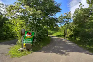 Eingang zum Campingplatz mit Schildern an der Straße, umgeben von Bäumen und grünem Hang.