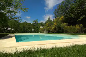 Türkisfarbenes Schwimmbecken mit Liegefläche am Rand, umgeben von Gras und Bäumen vor blauem Himmel.