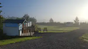 Campingplatzweg am Rasen entlang, neben einem kleinen Gebäude mit Holzgeländer und Schuppen, im Hintergrund Bäume und Nebel.