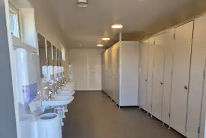 Reihe weißer Toiletten und Waschbecken in einem hellen Sanitärraum mit Kabinen und Fliesenboden.