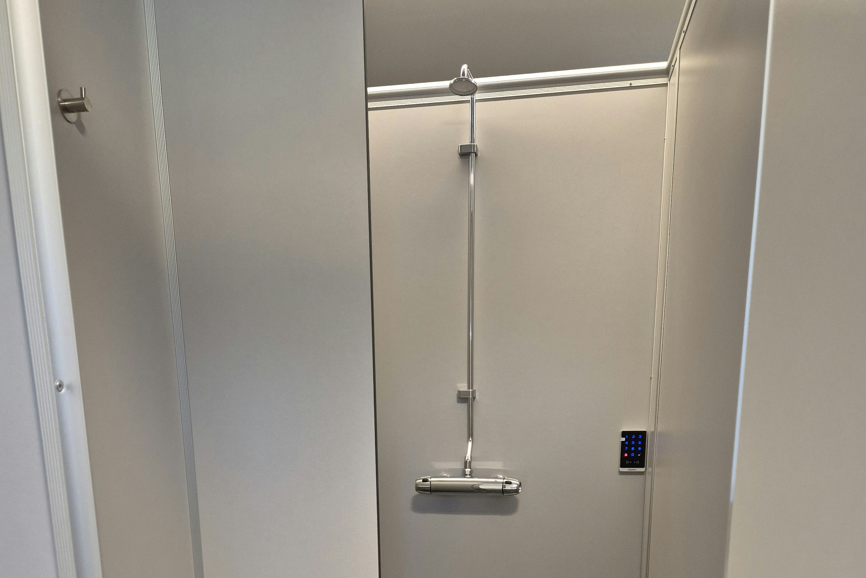 Barrierefreie Duschkabine mit Duschkopf, Haltegriffen und grauen Fliesen, daneben eine Ablage und eine Tür.