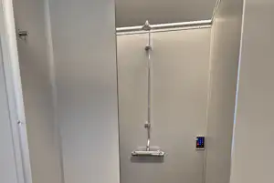 Barrierefreie Duschkabine mit Duschkopf, Haltegriffen und grauen Fliesen, daneben eine Ablage und eine Tür.