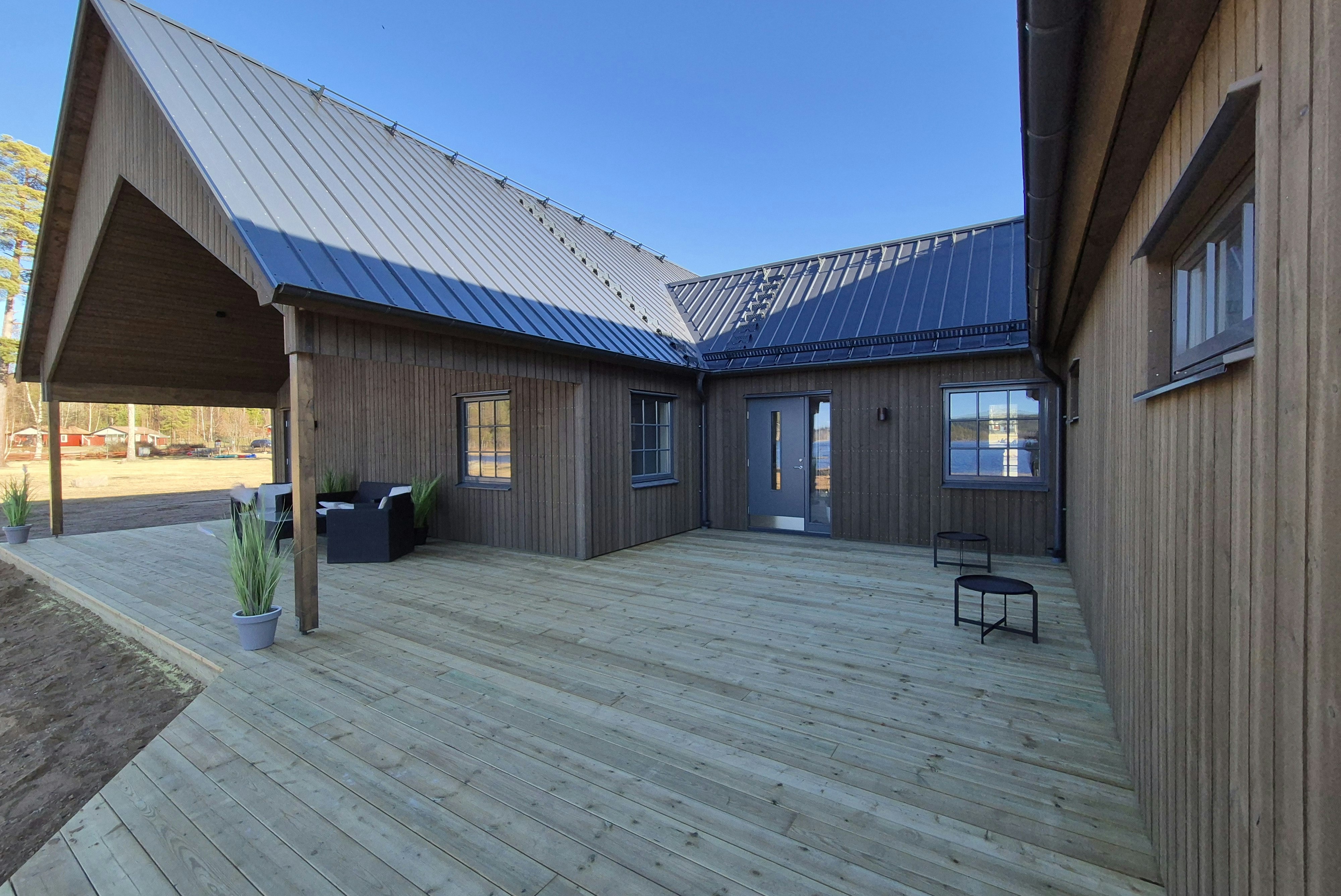 Holzgebäude mit überdachter Terrasse, grauem Boden und Sitzbank, daneben Mülltonnen und Fensterfronten unter blauem Himmel.