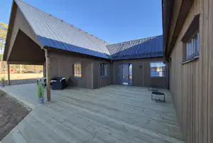 Holzgebäude mit überdachter Terrasse, grauem Boden und Sitzbank, daneben Mülltonnen und Fensterfronten unter blauem Himmel.