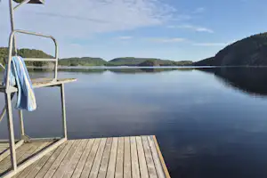 Seeterrasse am Steg mit Blick auf ruhiges Wasser und bewaldete Ufer, im Hintergrund ein See und Hügel.