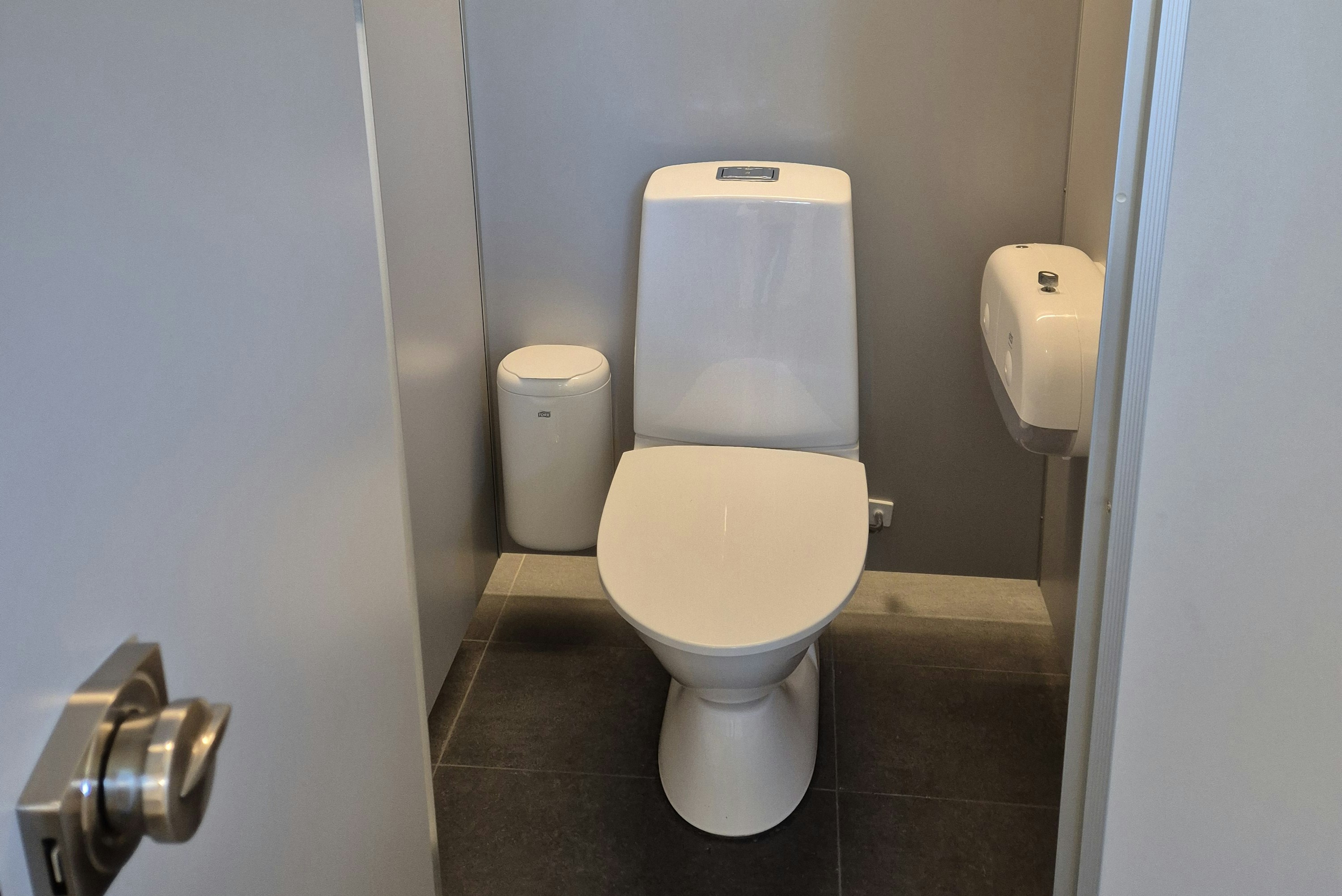 Weiße Toilette im modernen Badezimmer mit Fliesenboden und geschlossener Tür, seitlich ein kleines Waschbecken.