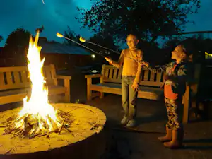 Lagerfeuer am Campingplatz mit Holzscheiten, daneben Sitzbänke und zwei Kinder, die am Abend am Feuer stehen.