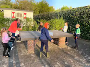 Kinder spielen an einer Tischtennisplatte auf dem Campingplatz, im Hintergrund ein Holzbungalow und Hecken im Garten.